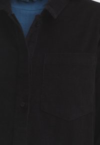 Čierna kordurová košeľa s vreckom, zapínaním na gombíky a špicatým golierom. Tkanina s textúrou a viditeľnými ryhami, čiastočne zakrytá modrou vrstvou pod ňou.