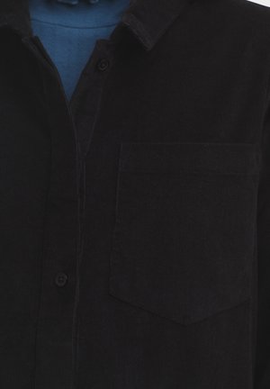 Zwarte corduroy overhemd met een zak, knoopsluiting en een puntkraag. Getextureerde stof met zichtbare ribbels, gedeeltelijk bedekt door een blauwe laag eronder.