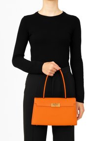 Oranje leren handtas met een gestructureerde vorm, voorzien van een gouden sluiting en dubbele handvaten, vastgehouden in de hand van een persoon tegen een witte achtergrond.