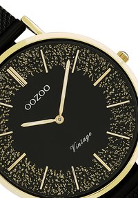 Montre noire avec accents dorés, cadran rond, aiguille noire texturée avec des points dorés et des index horaires dorés. Dispose d'un bracelet en maille et d'une couronne.