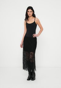 Black tank top combined with a sheer black lace maxi skirt. The skirt features a high slit, revealing a layered, textured design. Black ankle boots.