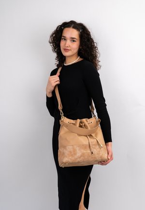 Borsa a secchiello in pelle color tan con una finitura morbida, tracolla regolabile, chiusura con cordoncino e dettagli in ottone. Superficie liscia e testurizzata.