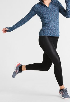 Mujer con camiseta de manga larga azul y leggings negros saltando a medio aire, lleva zapatos deportivos grises y rosas sobre un fondo liso.