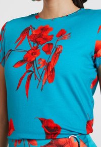 Top de manga corta en turquesa con un patrón floral en rojo, con gráficos llamativos de flores y hojas en toda la tela. Textura suave.