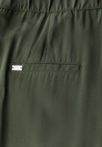 Olijfgroene stof met een gladde textuur, voorzien van een horizontale zak met een zilveren hardware logo-label aan de rechterkant in het midden.
