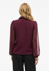 Chemisier bordeaux à motif à pois, avec des manches longues transparentes et un col classique. Texture lisse avec des poignets de manches froncés.