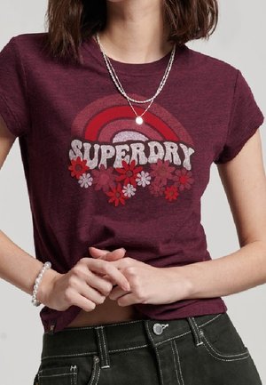 Femme portant un t-shirt Superdry bordeaux avec un motif arc-en-ciel et fleurs rouges, associé à un pantalon foncé, un collier en argent et un bracelet de perles.