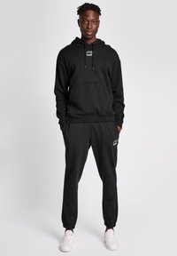 Svart huvtröja med framficka och dragsko, i kombination med matchande joggerbyxor med logotypdetaljer. Bärs med vita sneakers.