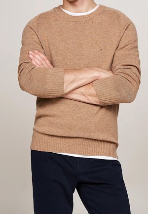 Homme bras croisés portant un pull en maille beige sur une chemise blanche et un pantalon bleu marine foncé sur un fond beige uni.