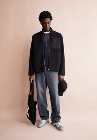 Dunkelgraue Fleecejacke mit einer Taschenpatte, über einem marineblauen Cardigan getragen; kombiniert mit grauen Jeans und leichtgewichtigen, mehrfarbigen Sneakers; trägt eine schwarze Handtasche.