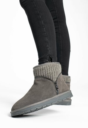 Botín de ante gris con un puño de punto gris, cremallera lateral y suela de goma texturizada. Combinado con jeans ajustados negros.
