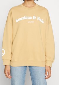 Sudadera beige con texto blanco "Sunshine & Rain", cuello redondo, hombros caídos y detalle de símbolo de paz en la manga. Ajuste suave y relajado.