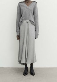 Maglione grigio lavorato a maglia con scollo a V e maniche ampie, abbinato a una gonna grigia fluida con cintura e orlo asimmetrico, indossato con stivali neri alla caviglia.