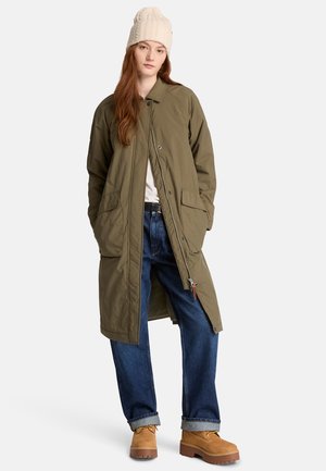 Vrouw draagt een lange olijfgroene jas, blauwe spijkerbroek, beige laarzen, wit overhemd en een crèmegebreide muts, staande tegen een effen achtergrond.