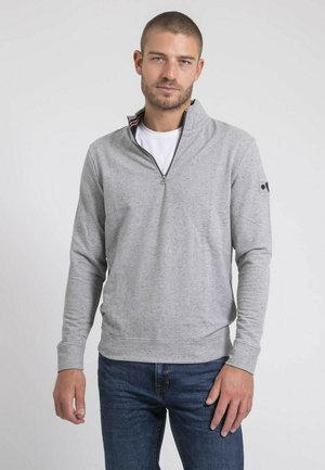 Homme portant un sweat-shirt gris clair à demi-fermeture éclair par-dessus une chemise blanche et un jean bleu, debout devant un fond clair uni.