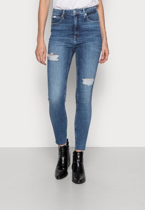 Pantalones ajustados de mezclilla azul de tiro alto con detalles desgastados, que presentan desgarros en la parte frontal y un acabado suave, usados con botas negras hasta el tobillo.
