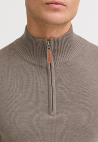 Suéter marrón con cremallera y cuello alto acanalado, con una cremallera de metal y una lengüeta de cuero con la etiqueta "BLEND". Textura de punto suave.