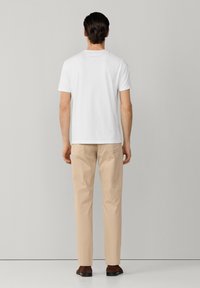 Mann mit dunklen Haaren, der ein schlichtes weißes T-Shirt, beige Hosen und braune Schuhe trägt und einem hellgrauen Wand gegenübersteht.