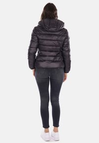 Giacca nera puffa con cappuccio, caratterizzata da sezioni orizzontali e una texture lucida. Abbinata a jeans scuri e sneakers bianche.