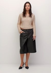 Femeie cu părul ondulat și închis la culoare, îmbrăcată într-un pulover bej pufos, pantaloni culotte din piele negri cu croi larg și pantofi cu vârf ascuțit negri, stând pe un fundal simplu.