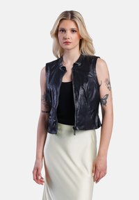 Černá kožená zkrácená vesta s lesklou texturou, se zipem a bez rukávů. Nositá přes přiléhavé černé tričko a světlou sukni.