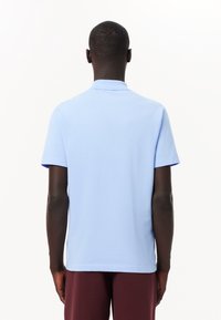 Homme vu de dos portant un polo à manches courtes bleu clair et un pantalon rouge foncé sur un fond blanc uni.