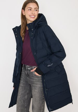 Leende kvinna klädd i en marinblå lång vadderad vinterjacka med huva, lila tröja och bleka gråjeans, med handen i jackfickan.
