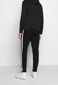 En person i en svart hoodie och svarta joggers med vita sidstrimlor ses bakifrån, stående mot en neutral bakgrund.