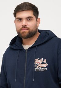 Navy blue zip-up hoodie with a drawstring hood, featuring a white and red logo on the chest, made from soft fabric.