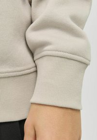Beige sweatshirtmudd med ribbad textur och en tajt passform. Sömmar är synliga på kanten av muddarna. Handen vilar försiktigt nedanför.
