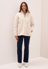 Veste en polaire beige avec col montant, fermeture éclair à l'avant, et poche poitrine. Portée avec un jean bleu foncé et des baskets blanches. Tissu à texture lisse.