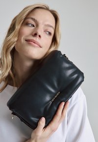 Zwarte leren clutch met een zachte textuur, ritsluiting en een goudkleurig reliëflogo. Compacte rechthoekige vorm, ideaal voor handmatig gebruik.