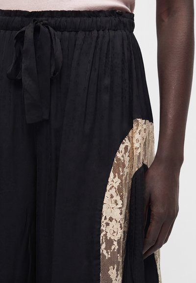 Μαύρη φούστα με μπεζ λουλουδάτη δαντελωτή επένδυση, ελαστική μέση και δεμένη κορδέλα, φορεμένη από άτομο με σκούρο δέρμα και ανοιχτό ροζ μπλουζάκι.