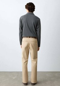 Bărbat cu păr închis la culoare, purtând o cămașă cu mânecă lungă gri închis, pantaloni bej, curea maro și pantofi maro, stând cu fața spre un perete alb.