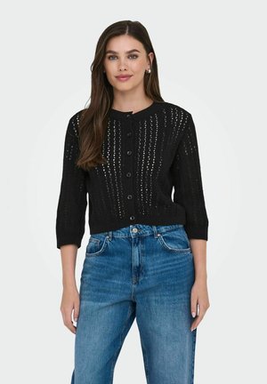 Cardigan noir en tricot avec un col rond, doté de fermetures à boutons et d'un motif texturé. Porté sur un jean bleu taille haute.