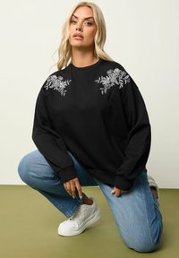 Felpa nera con ricami floreali bianchi sulle spalle, maniche lunghe, collo rotondo e vestibilità rilassata. Indossata con jeans azzurri chiari e sneakers bianche.