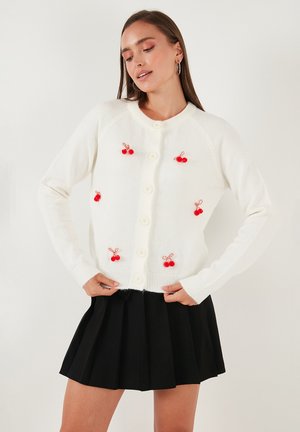 Valge kardigan punaste kirsi kaunistustega, ümar kaelus ja nööbitav esiosa. Seljas mustal plisseeritud seelikut. Pehme tekstuur.