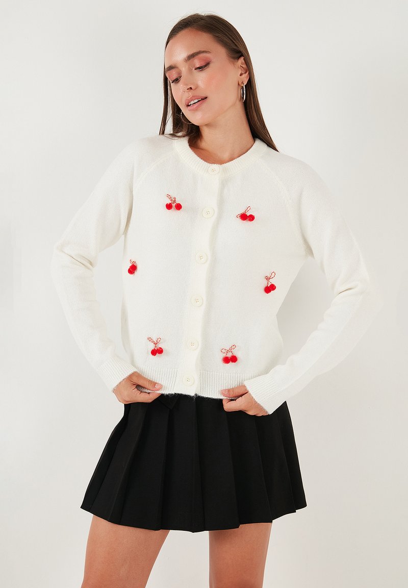 Witte cardigan met rode kersenversieringen, ronde halslijn en knoopsluiting aan de voorkant. Draagt over een zwarte geplooide rok. Zachte textuur.