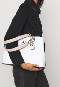 Hvid syntetisk håndtaske med præget logo, lyserøde stropper, sølvhardware og et aftageligt mærke, holdt under armen.