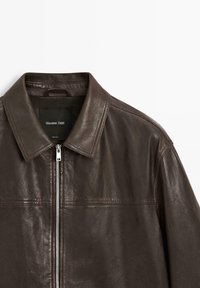 Dunkelbraune Lederjacke mit Reißverschluss und spitzem Kragen, flach auf weißem Hintergrund liegend.