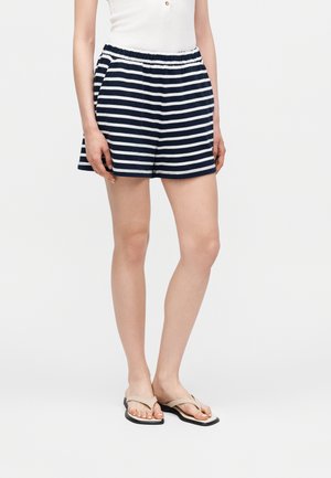 Femeie care poartă pantaloni scurți cu dungi orizontale bleumarin și albe, un top alb și sandale bej tip flip-flop, stând într-un fundal simplu.