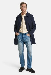 Marineblauwe katoenen trenchcoat, aan de voorkant knopen, over een lichtgestreept overhemd, gecombineerd met vervaagde blauwe spijkerbroeken en donkere schoenen.