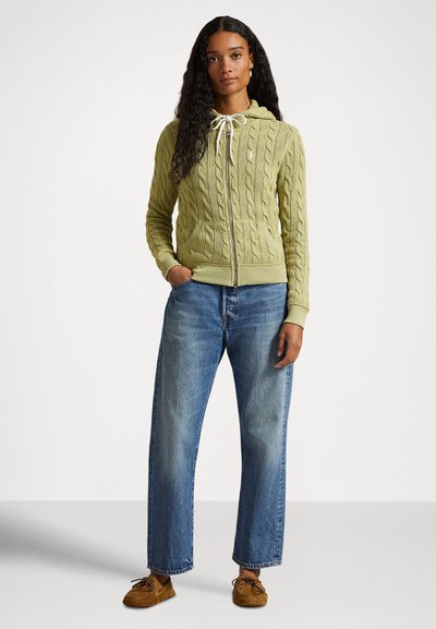 Jauna moteris su ilgais garbanotais plaukais, vilkinti šviesiai žalią megztą užtraukiamą džemperį, mėlynas džinsas ir ruda mokasinų avalynė, stovinti su rankomis kišenėse.