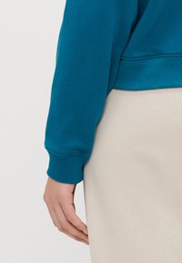 Felpa color teal con polsini a costine, mostrata insieme a un capo di colore crema. Tessuto dalla consistenza liscia, vestibilità rilassata e design semplice.
