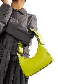 Borsa a spalla in pelle giallo neon con forma curva, tracolla regolabile e charm in metallo. Indossata su un indumento grigio scuro.