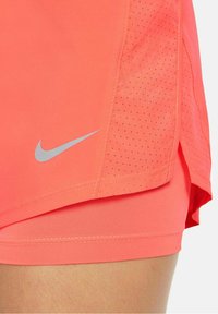 Pantalones cortos Nike en un vibrante color coral con un logo plateado, que cuentan con un panel de malla transpirable y una abertura lateral para una mejor movilidad. Textura suave.