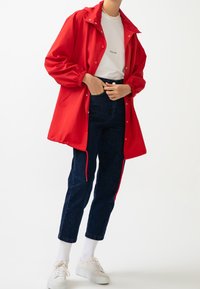Rød vandtæt jakke med trykknapper, elastiske manchetter, iført over en hvid T-shirt og mørkeblå jeans, parret med hvide sneakers.