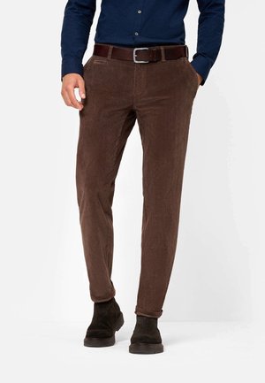 Mann trägt braune Cordhose mit umgeschlagenen Bündchen, dunkelbraunen Gürtel, dunkelblaues Hemd und dunkelbraune Wildlederstiefel, steht mit Händen in den Taschen.