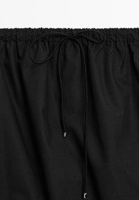 Cintura in tessuto nero con elastico e chiusura con cordoncino. Presenta punte metalliche sul cordoncino e una texture liscia e opaca.