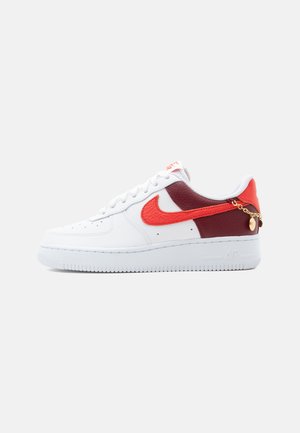 Baskets blanches Nike Air Force 1 avec des panneaux rouges et grenat, un swoosh rouge, et une petite chaîne en or attachée près du talon.